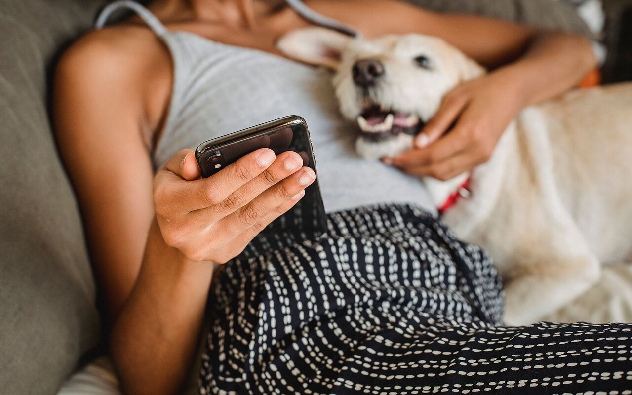 Tutora en el sillón con el teléfono en la mano y su perro recostado al lado.